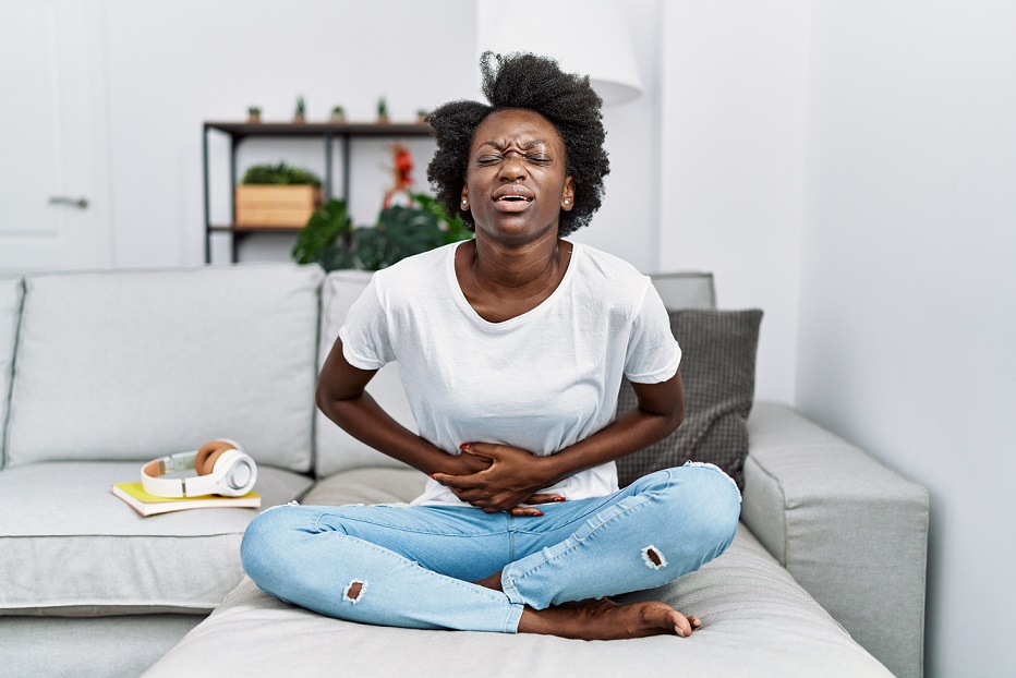 African,Young,Woman,Sitting,On,The,Sofa,At,Home,With hands gripping her belly in pain. depicting Symptom of fibroid