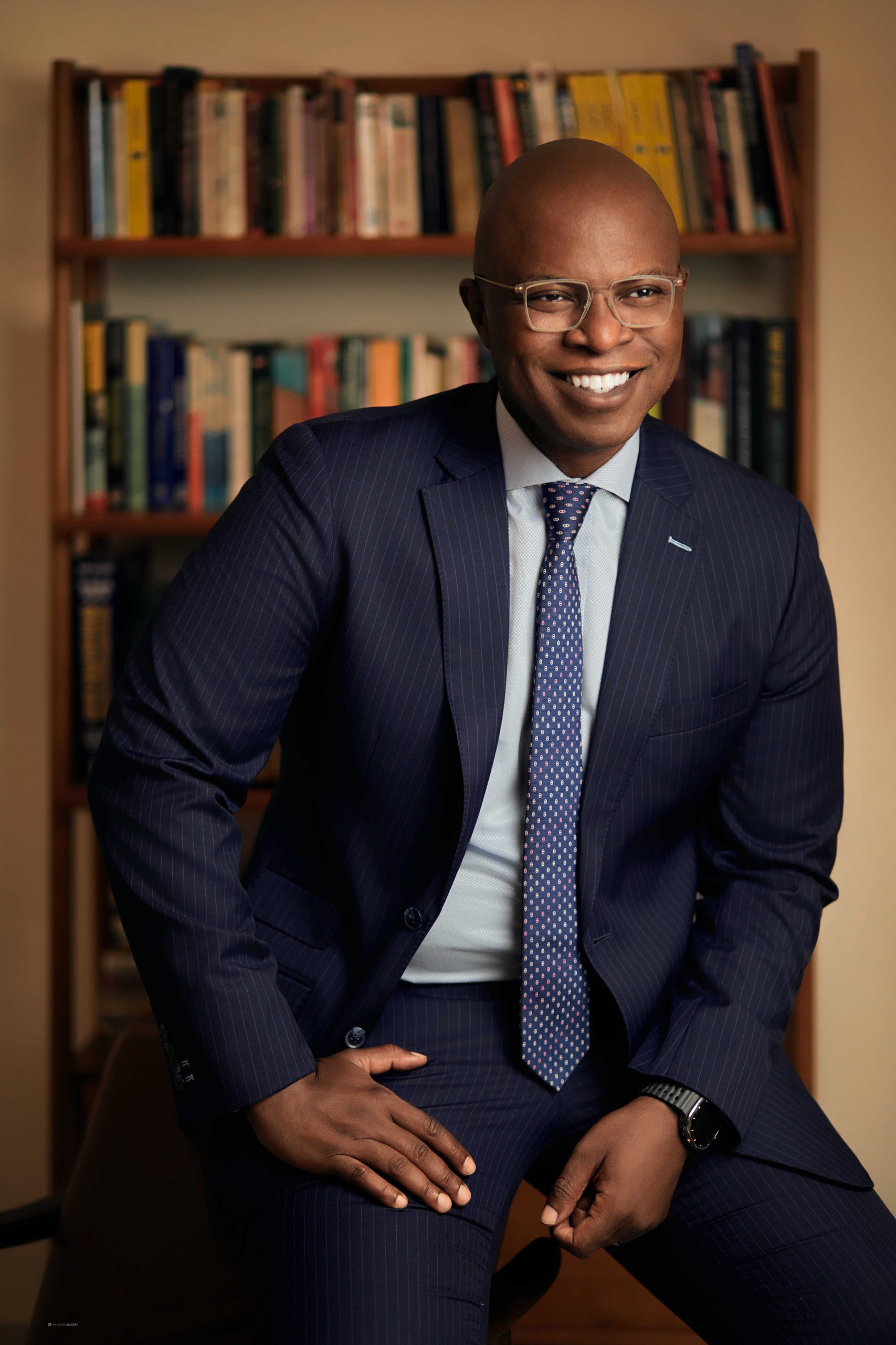 Portrait of Sim Shagaya, smiling in a navy blue pinstripe suit with a white shirt and patterned tie, sitting in front of a bookshelf. He wears glasses and a wristwatch, with one hand resting on his knee.