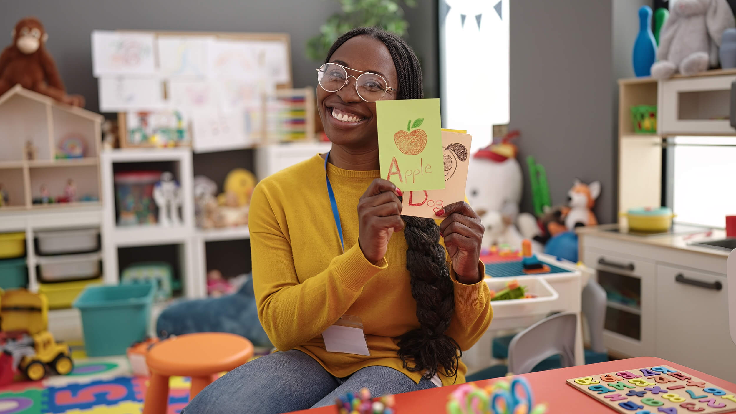 Smiling female teacher in glasses and yellow sweater sitting in a colorful classroom, holding up a flashcard with the word ‘Apple’ and letter ‘A’, with toys and learning materials in the background.