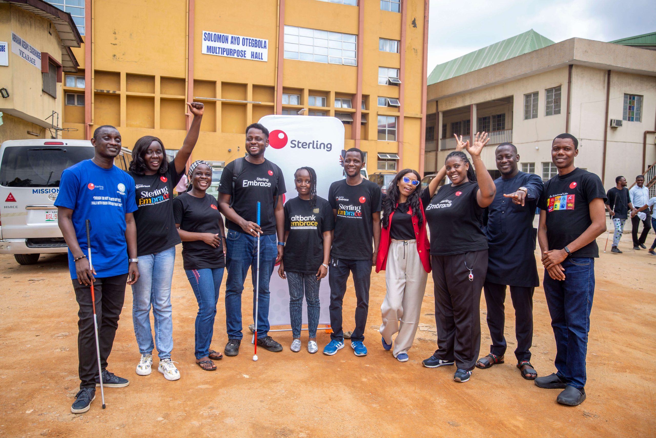 Group of smiling people posing outdoors in front of a yellow building during a Sterling Bank community outreach event for the visually impaired, with participants wearing branded “Sterling Embrace” and “AltBank Embrace” shirts.