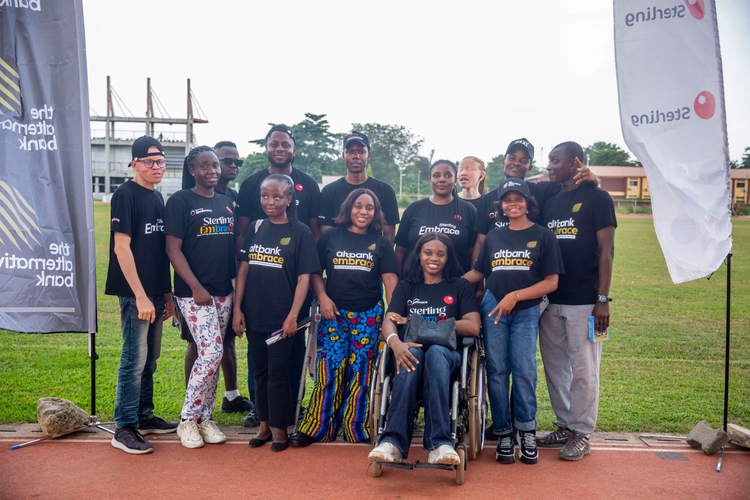 A group of smiling participants wearing Sterling Bank and AltBank Embrace t-shirts posing together outdoors on a sports field. The group includes individuals standing and one person seated in a wheelchair, with Sterling-branded flags visible on both sides.