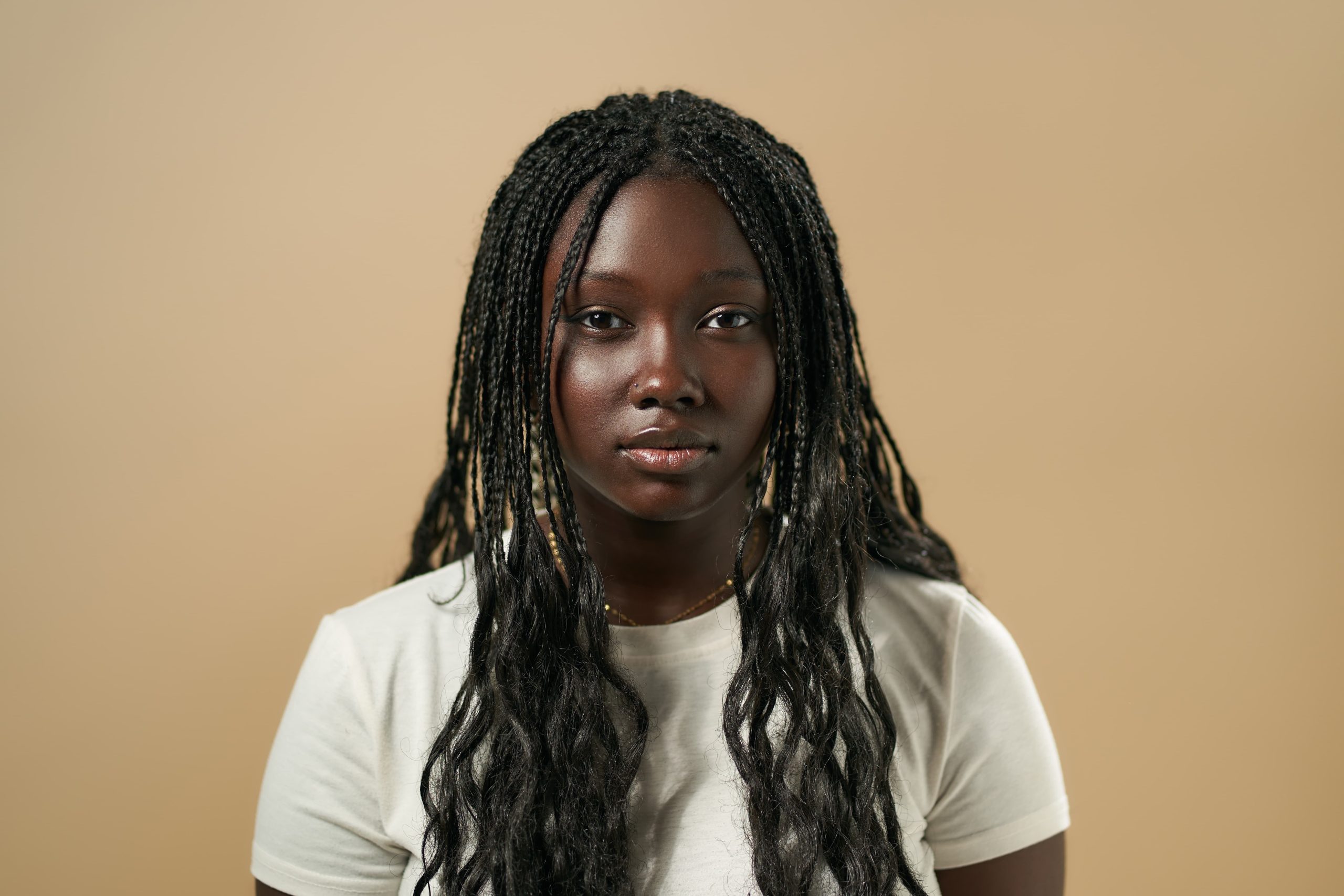 Portrait of a young African woman with long braided hair wearing a white t-shirt against a neutral beige background, looking calmly at the camera.