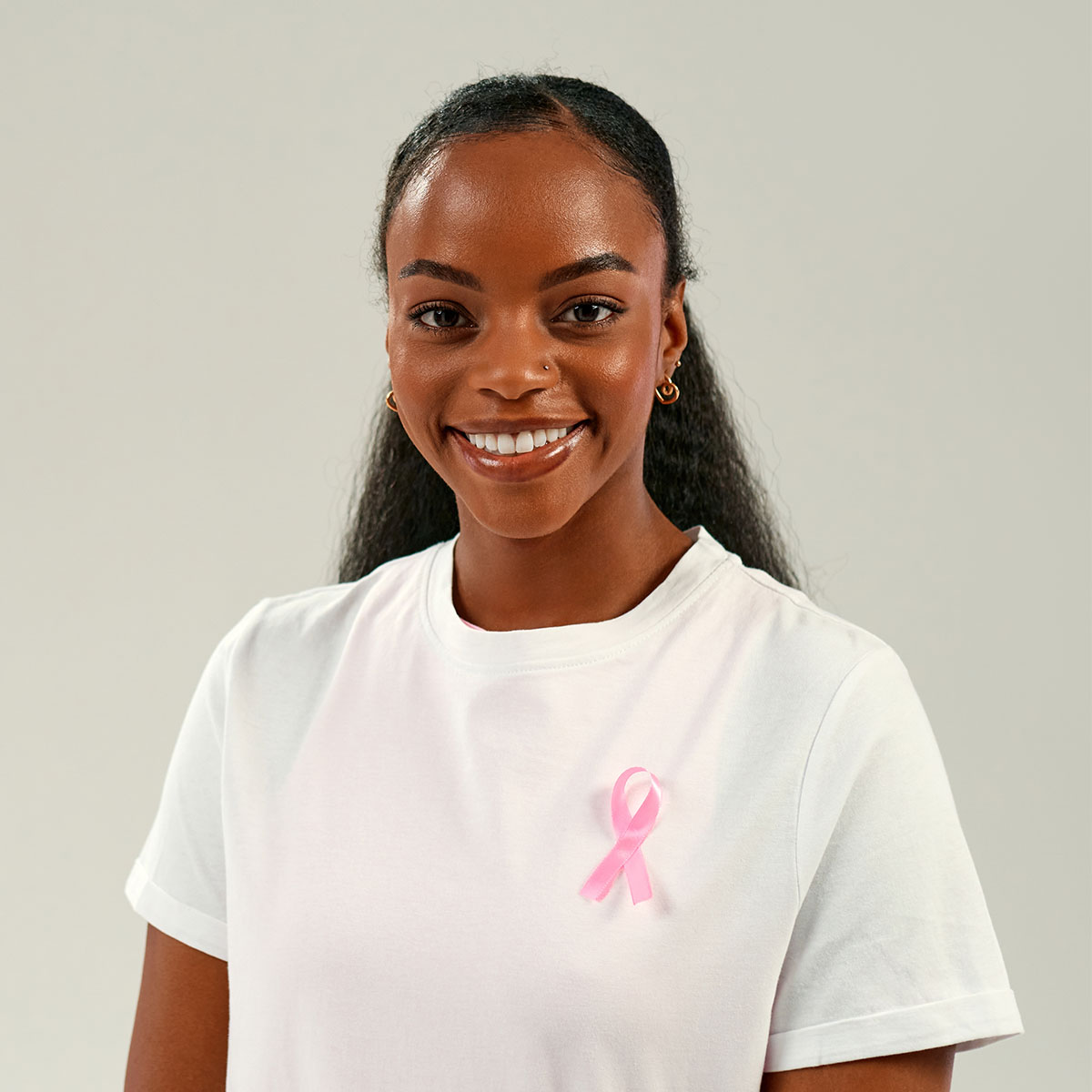 Smiling young woman wearing a white t-shirt with a pink cancer awareness ribbon pinned to it, standing against a neutral background.