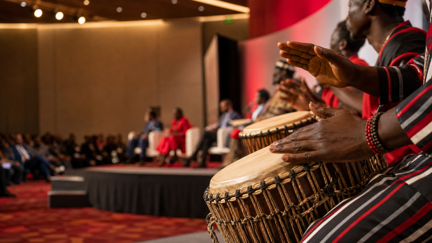 African drummers perform at a social impact summit, with a blurred audience and panel session in the background.